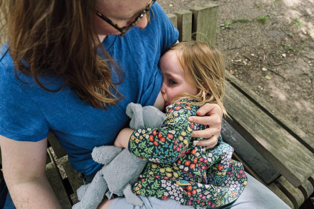 Tired child breastfeeding while held on bench by mum during a documentary family photo session